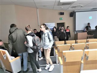 Uczestnicy z plecakami rozmawiają między rzędami sali wykładowej.