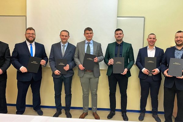 Grupa absolwentów prezentuje oprawione prace dyplomowe w sali uczelni.