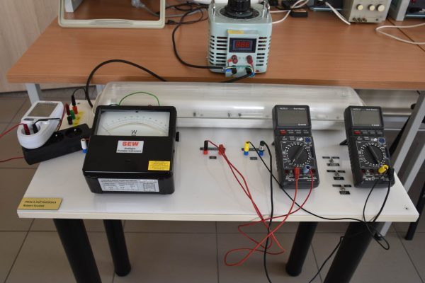 Stanowisko laboratoryjne z miernikiem analogowym, multimetrem i połączonym przewodami układem.