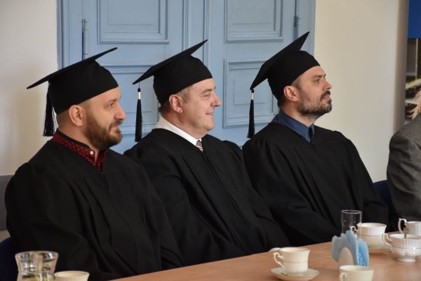 Trzej absolwenci w togach i biretach siedzą przy stole podczas uroczystości.