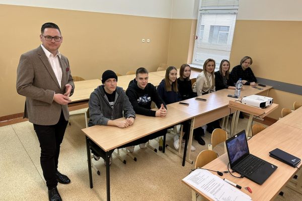 7 młodych osob siedzi przy ławkach i uśmiecha się. Tuz obok stoi mężczyzna, który również się usmiecha