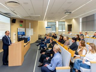 Mężczyzna stoi przy mównicy w sali wykładowej i przemawia do zebranych osób