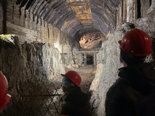 Uczestnicy w kaskach oglądają sklepienie podziemnego korytarza ze stalowymi podporami.
