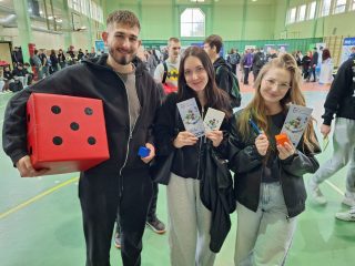Troje uczestników targów edukacyjnych prezentuje ulotki i dużą czerwoną kostkę do gry.
