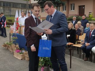 Wręczenie dyplomu i upominku uczniowi podczas uroczystości na szkolnym dziedzińcu.