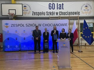 Przemówienie przy mównicy w Zespole Szkół w Chocianowie, obok flagi Polski i Unii Europejskiej.