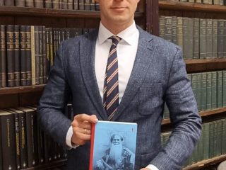 Mężczyzna prezentuje książkę na tle bibliotecznych regałów.