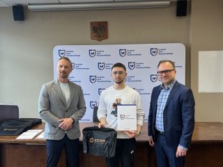 Mężczyzna w białej koszulce prezentuje certyfikat KGHM i torbę upominkową.
