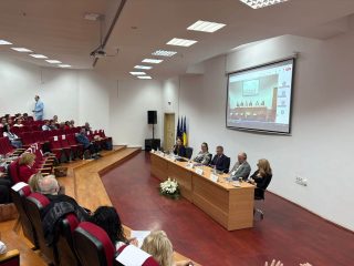 Obrady konferencji w auli z uczestnikami przy stole i połączeniem online na ekranie.