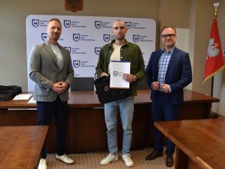 Mężczyzna w zielonej koszuli prezentuje certyfikat i torbę upominkową KGHM.