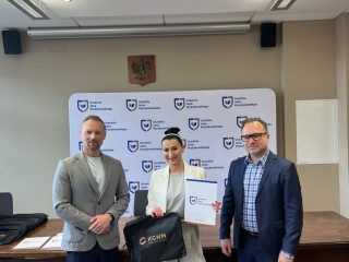 Wyróżniona w jasnym garniturze prezentuje certyfikat i torbę KGHM pomiędzy wręczającymi.