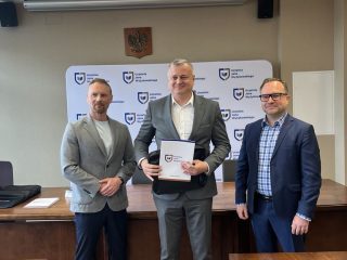 Mężczyzna w okularach i szarej marynarce pozuje z certyfikatem pomiędzy wręczającymi.