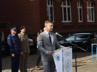 Mężczyzna w szarym garniturze przemawia przy mównicy na szkolnym dziedzińcu.