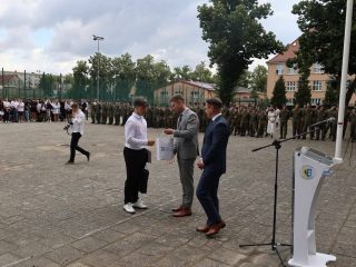 Uczeń odbiera dokument na placu przed szkołą w obecności dwóch mężczyzn.