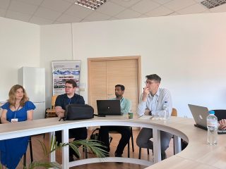 Uczestnicy konferencji siedzą przy zakrzywionym stole z laptopami.