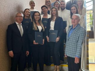 Grupa absolwentów i przedstawicieli uczelni pozuje na schodach z żółtymi oznaczeniami.