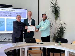 trzech uśmiechniętych mężczyzn stojących przy stole i wspólnie trzymających certyfikat