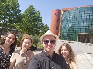 Grupowe selfie czworga uczestników przed budynkiem kampusu.