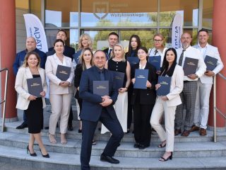 Absolwenci prezentują oprawione prace na schodach przed UJW; mężczyzna z przodu pozuje z szeroko rozstawionymi nogami.