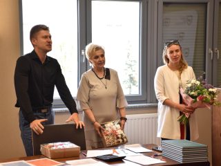 Troje członków komisji stoi za stołem z pracami; kobieta trzyma bukiet kwiatów.