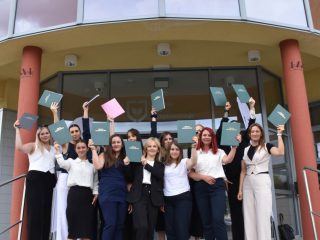 Grupa absolwentek unosi prace dyplomowe na schodach przed uczelnią.