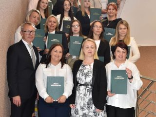 Uczestnicy obron zarządzania pozują na schodach wewnątrz uczelni z zielonymi pracami dyplomowymi.