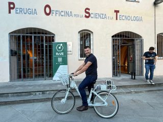 Mężczyzna na rowerze przed centrum nauki Perugia Officina per la Scienza e la Tecnologia, obok banera H2VE.