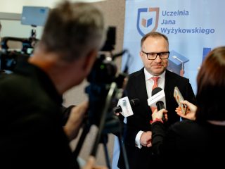 Mężczyzna w okularach udziela wywiadu dziennikarzom z mikrofonami przed kamerą.