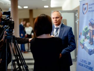 Mężczyzna w niebieskim garniturze udziela wywiadu przy banerze UJW.
