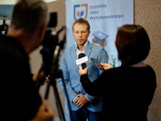 Mężczyzna w jasnoniebieskim garniturze rozmawia z dziennikarką przed kamerą.