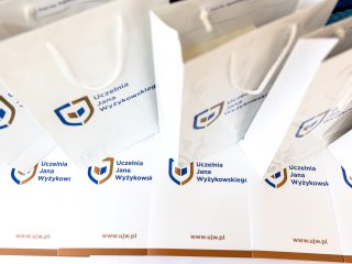 Białe torby upominkowe i dokumenty z logo Uczelni Jana Wyżykowskiego.