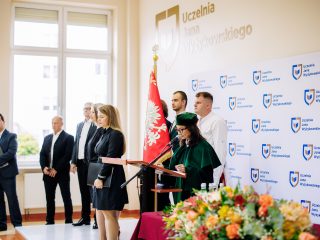 Uczestnicy inauguracji stoją w auli, a osoba w stroju akademickim przemawia przy mównicy.