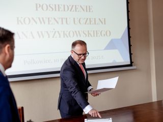 Mężczyzna w okularach trzyma otwartą teczkę przy stole konferencyjnym.