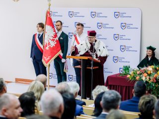 Widok auli podczas inauguracji z pocztem sztandarowym i władzami uczelni na podium.