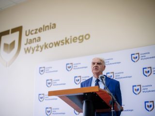 Mężczyzna w niebieskim garniturze przemawia przy mównicy pod nazwą uczelni.