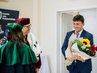 Uśmiechnięty mężczyzna z bukietem kwiatów rozmawia z przedstawicielami uczelni w strojach akademickich.