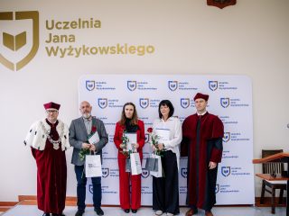 Trzy wyróżnione osoby z kwiatami i upominkami pozują pomiędzy przedstawicielami uczelni w togach.