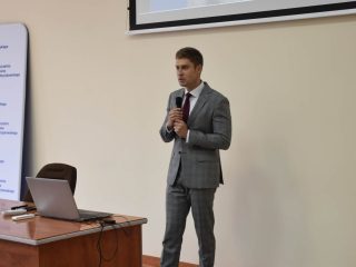 Prelegent w szarym garniturze przemawia na tle slajdu z historyczną fotografią.