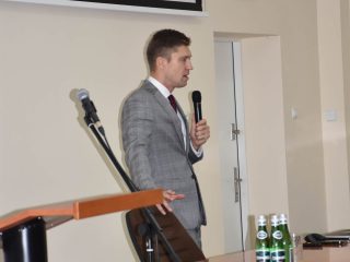 Mężczyzna w szarym garniturze prowadzi wykład z mikrofonem przy stole.