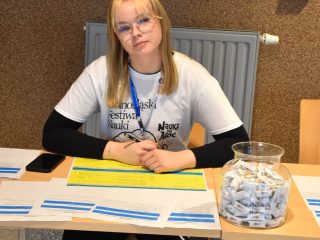 Kobieta przy stoliku rejestracyjnym festiwalu nauki z formularzami i naczyniem cukierków.