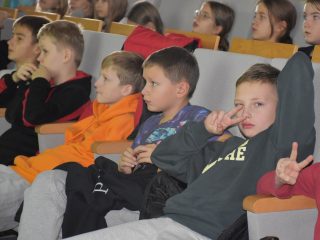 Dzieci w pierwszym rzędzie podczas wykładu; chłopiec pokazuje gest zwycięstwa.