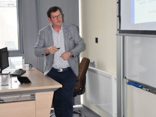 Prowadzący w jasnoszarej marynarce stoi przy ekranie w sali ćwiczeniowej.