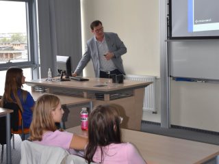 Prowadzący przy komputerze prezentuje materiały uczniom siedzącym w ławkach.