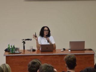 Prelegentka przy laptopie prowadzi wykład przed uczniami w auli.
