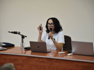 Prelegentka gestykuluje podczas wykładu, siedząc za stołem.
