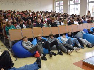 Widok pełnej auli; część uczestników siedzi na niebieskich pufach przed pierwszym rzędem.
