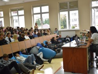 Prelegentka prowadzi wykład przed publicznością siedzącą w auli i na pufach.