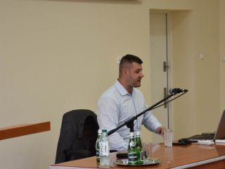 Prelegent przy mikrofonie i laptopie podczas wykładu w auli UJW.