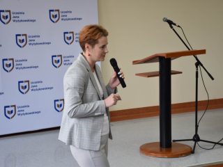 Kobieta w jasnym garniturze przemawia do mikrofonu na tle logo UJW.