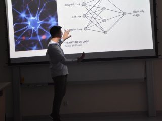 Prelegent wskazuje schemat sieci neuronowej na slajdzie „Neuron i sieć”.